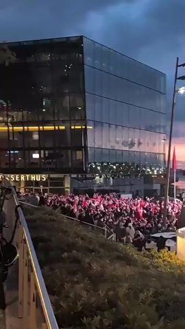 A large crowd of people, mostly wearing red and white, are gathered outside a modern glass-fronted building. The sky above is a deep blue with hints of pink and orange from a sunset. Red flags are visible among the crowd and in front of the building. To the right, there is a stage with a large screen and scaffolding, and several white tents with illuminated interiors. People are standing and milling around, some appearing to be watching something on the screen. The overall atmosphere appears to be festive.