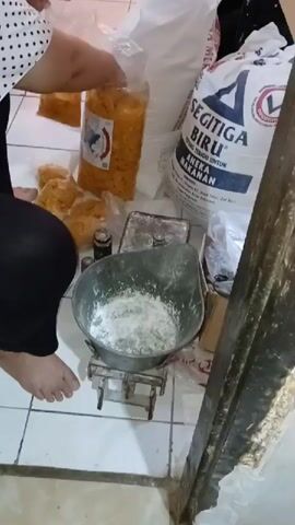 A person's feet and lower legs are visible on the left side of the frame. They are wearing black pants. The person is interacting with a large clear plastic bag filled with yellow, crunchy snack items. Next to this bag, there are several smaller clear plastic bags, also containing the same yellow snack items. In the center of the frame, there is a vintage metal scale with a large metal bowl on top. The bowl contains a white powdery substance, likely flour. Behind the scale, there are two small weights. To the right of the scale, a large white bag with blue and red lettering is visible. The text on the bag appears to be in Indonesian and includes the words "SEGITIGA BIRU" and "TEPUNG TERIGU UNTUK ANEKA MAKANAN". This indicates the bag contains wheat flour for various foods. The environment appears to be an indoor space with tiled flooring. The overall scene suggests food preparation or packaging.