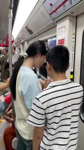 The video shows a subway car in what appears to be China. Several passengers are visible. A young woman with long dark hair, wearing a light blue and tan shirt and jeans, stands facing a young boy in a striped shirt. The boy is looking up at the woman, and she appears to be adjusting something near his face, possibly his glasses. Other passengers are in the background, some looking at their phones. There are various handrails and signage visible inside the subway car.