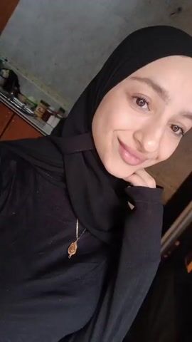 A woman in a black hijab and black top smiles slightly at the camera, her chin resting on her fist. She wears a gold necklace. She is in a kitchen with tile walls, cabinets, a stove, and miscellaneous items on the counter. Background music plays.
