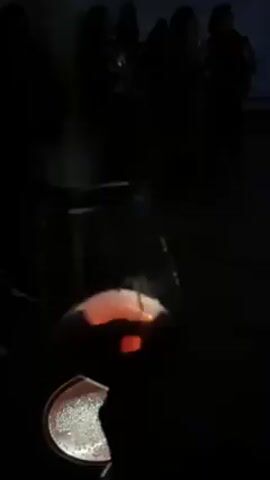 The video shows a dimly lit room with people standing in the background and a wine glass in the foreground. The camera focuses on the wine glass, which is filled with a dark liquid and has bubbles.  Behind the glass, there is a wall with white cylinders stacked in a grid pattern, some with pink glowing centers, and a tree branch projection above them. The lighting is subdued, focusing attention on the wine glass and the wall installation. The overall setting appears to be an art installation or exhibition.
