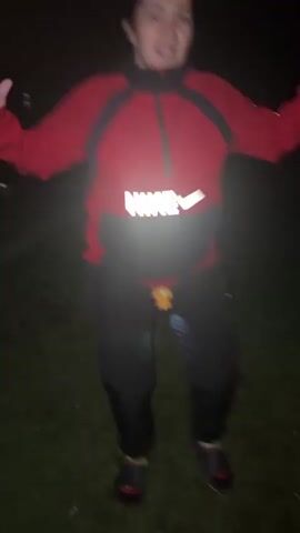 A woman in a red Nike hoodie and black pants is standing in a grassy area at night. She is waving her hands and appears to be talking. There is a small yellow object attached to her pants.