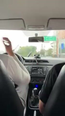 The video shows a view from the back seat of a car as it drives through a city on a rainy day. The driver is wearing a white shirt and adjusts the car's radio or climate control. The passenger in the front seat is wearing a black shirt. Through the windshield, other cars and buildings are visible. The car's interior is visible, including the dashboard, steering wheel, and rearview mirror.