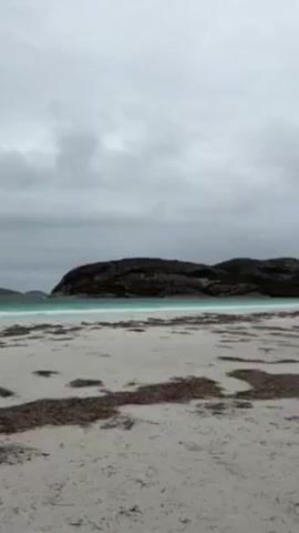 The video shows a beach on an overcast day. The sand is light-colored and scattered with seaweed. The water is a striking turquoise color, and there are gentle waves rolling in. In the distance, there are rocky, island-like formations that break up the horizon. The sky is covered with grey clouds.