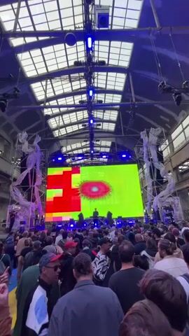 The video shows a crowded concert venue. A large LED screen displays colorful, abstract graphics, accompanied by a light show. The venue is a large industrial space with high ceilings and metal beams, with a latticework of skylights.  A crowd is facing the stage, some individuals looking at their phones. There are supporting structures to the side of the LED screen, draped with white material.