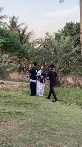 This media file shows three individuals in an outdoor setting. Two people, a man and a woman, are standing together in the center, facing the camera. The man is wearing a dark blue traditional outfit with white accents, and the woman is wearing a white and black patterned dress and a hijab. The woman is holding a bouquet of light blue and white flowers. To the right of them, a third person, a man, is standing and appears to be directing them or perhaps taking a photograph with a phone. He is wearing a black t-shirt and dark pants. The scene is set on a grassy area with a brick wall and lush greenery, including palm trees and other tropical vegetation, in the background. The sky suggests it is late afternoon or early evening, with soft, diffused light. The overall atmosphere appears to be a casual photoshoot or a celebratory moment, possibly an engagement or wedding. The location appears to be Kembang Harum, Indonesia, given the tropical flora. There is no visible text in the media file.