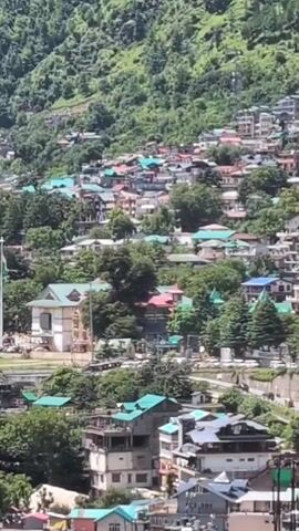 A drone shot pans across a densely populated hillside town. The houses are multi-story, with a variety of colored roofs, including teal, red, and gray. They are nestled amongst lush green trees. In the background, a steep, tree-covered mountain rises under a clear blue sky. The town appears to be built on a significant slope, with some buildings appearing to be constructed on stilts. A large, white building with a distinctive architectural style stands out on the left side of the frame, possibly a public building or a significant residence. The overall impression is one of a vibrant, lived-in community integrated into a natural, mountainous landscape. No people are visible.