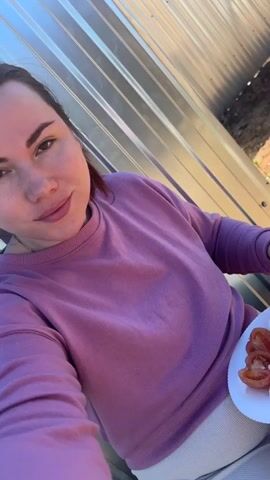 A woman in a purple sweatshirt is leaning against a corrugated metal wall outside, eating a dish with a spoon. She has her hair pulled back and is wearing a necklace. She takes a spoonful of food, closes her eyes and chews with a look of enjoyment on her face. She then opens her eyes and continues chewing with her lips pursed. There is a plate of sliced tomatoes visible in her lap. The background shows a bright blue sky.