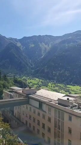 The video pans across a mountainous landscape on a clear day. In the foreground, the upper floors of a light-colored, multi-story building are visible, with some architectural details such as windows, pipes, and a walkway with railings. Beyond the building, a valley unfolds, dotted with trees and a small town with houses and buildings. The valley is surrounded by lush green mountainsides covered in dense trees, with rocky peaks in the distance. The sky is a clear blue with a few wispy clouds. The camera movement is smooth, simulating a slow panoramic sweep of the scenery.