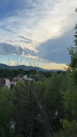 The video shows a time lapse of a landscape under a cloudy sky. The camera is positioned at a high angle, looking down over a village with houses and trees. In the foreground, there is a bare tree with branches reaching towards the sky. The background consists of rolling hills and mountains. The clouds in the sky are white and gray, with some patches of blue sky visible. The sun is setting or rising, casting a warm glow on the horizon. The video is filmed in portrait orientation.