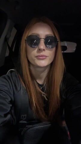 A woman with long red hair and sunglasses is sitting in a car at night. She is wearing a black top and a necklace. She is looking directly at the camera and speaking. The interior of the car is dark.
