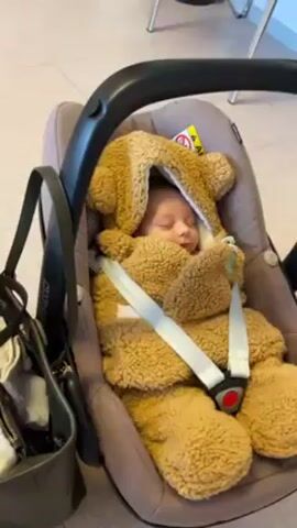 The video shows a baby sleeping in a car seat. The baby is wrapped in a fuzzy brown bear-shaped blanket. The car seat is brown and has a black handle. A black bag hangs next to the car seat. The background shows a light-colored tiled floor and a light blue wall with white chairs. The camera zooms in slightly on the baby's face. The baby makes a gurgling sound.