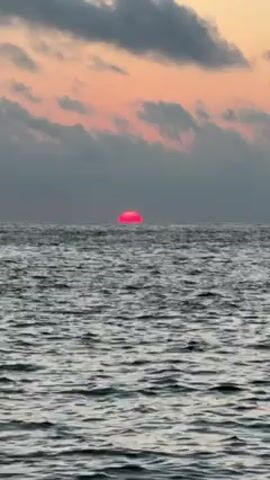 A video shows a pink sun setting over the ocean. The water is rippling, and there are gray clouds in the sky. The sunset is creating a pink and orange glow in the sky.