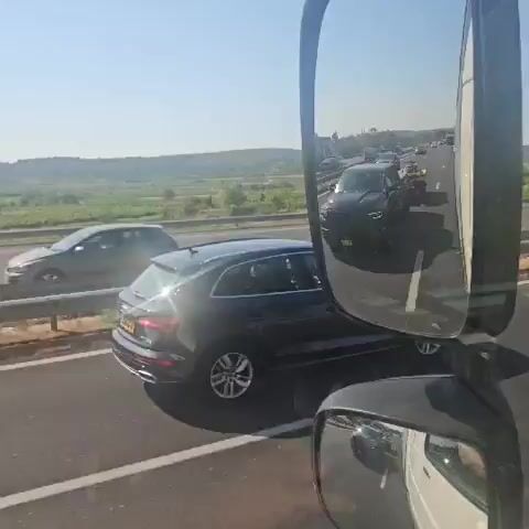The video is filmed from inside a vehicle, likely a truck, looking out through the passenger side mirror and windshield. The view shows a highway with traffic. In the foreground, the large passenger side mirror reflects the scene behind. Through the windshield, a black pickup truck is visible, towing a trailer with two yellow jet skis. The highway has multiple lanes, and other vehicles are driving in both directions. The environment appears to be a sunny day with a clear blue sky and rolling green hills in the background. The camera captures a sense of movement as the vehicle drives on the highway.
