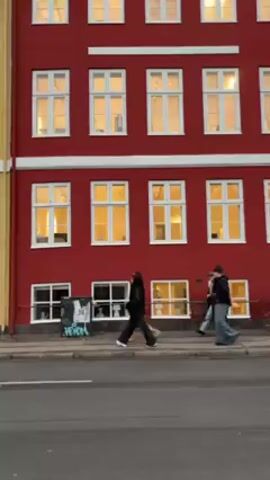 The video shows a three-story red building with white-framed windows. People are walking on the sidewalk in front of the building. A cyclist passes by in the foreground. The building is adjacent to a yellow building on the left. A street separates the building from the viewer.