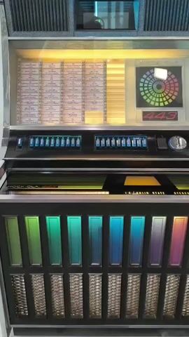 A close-up shot of a vintage Wurlitzer jukebox. The jukebox is silver and black with multicolored lights at the bottom. The selection panel is visible at the top, along with the song title display. The jukebox is in a brightly lit room, possibly a museum or a collector's home. The camera slowly zooms out, revealing more of the jukebox and its surroundings. The overall impression is one of nostalgia and retro charm.