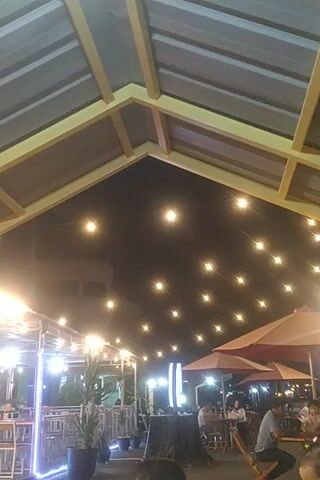 A low-angle, slightly tilted shot captures an outdoor dining area at night. The foreground is dominated by the corrugated metal roof and wooden beams of a structure, possibly a patio cover. A string of small, warm-toned lights is suspended across the dark sky, creating a festive atmosphere. In the background, several people are seated at tables, with some illuminated by additional lights from overhead fixtures and the string lights. Umbrellas are also visible, suggesting an outdoor seating arrangement. The overall impression is that of a casual dining establishment.