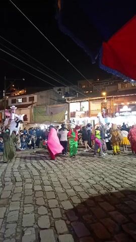The video shows a nighttime street celebration taking place on a cobblestone surface. A crowd of people is gathered, watching and participating in the event. Several individuals in colorful costumes are dancing and moving around. One person wears a bright pink outfit and twirls a cape, while another is dressed in green. There are also performers riding fake horses. Buildings line the street, and streetlights illuminate the scene. A red and blue banner is visible in the foreground. The overall atmosphere appears festive and lively.