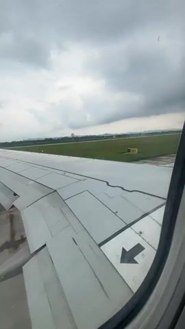 The video shows a view from an airplane window. The airplane is on the tarmac, possibly taxiing or preparing for takeoff. The wing of the airplane is prominently visible in the foreground, with various mechanical components and panels. The view outside the window reveals a runway and a grassy area beyond, with some distant structures and a cloudy sky. There are no people or interactions visible in the frame. The lighting suggests it is daytime. The perspective is fixed, looking out from the aircraft.