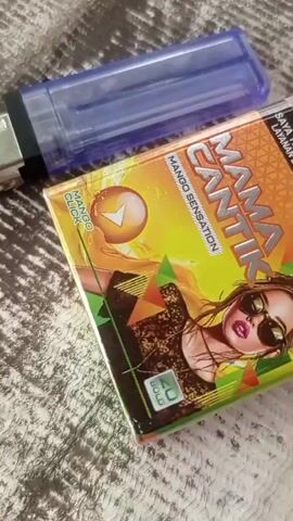 A close-up shot shows a pack of "Mama Cantik Mango Sensation" cigarettes next to a blue disposable lighter. The cigarette pack features a vibrant design with a woman wearing sunglasses and colorful geometric shapes. A label indicating "20 BOLD" is visible on the bottom right corner of the pack. The objects are placed on a textured surface, possibly a blanket or fabric. The lighting is soft, casting a slight shadow. The video is brief, with no discernible action other than the still image.