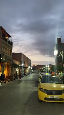 The video showcases a street scene in what appears to be a Middle Eastern or North African city during dusk or twilight. The street is lined with buildings, mostly two or three stories high, with visible architectural details like arched windows and balconies. The buildings are lit by decorative street lamps, casting a warm glow. On the right side of the street, several yellow taxis are parked. People are walking along the street, appearing as silhouettes or indistinct figures due to the low light. In the distance, the skyline is punctuated by a prominent dome and a tower-like structure, possibly part of a mosque or historical building, which is also illuminated. The sky is overcast with a mix of gray and soft orange hues, suggesting the transition from day to night.