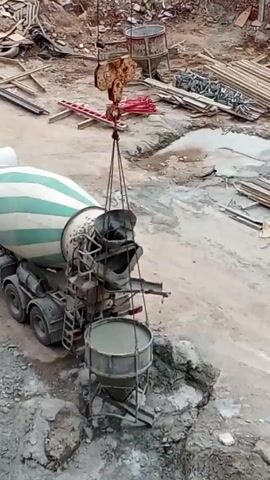 The scene is a construction site. A large, green and white striped cement truck is parked on the left. A crane is centrally positioned, lifting a metal bucket filled with cement. A construction worker in a yellow jacket and white hardhat walks towards the bucket. The ground is littered with construction debris, including metal bars, wooden planks, and other equipment. The overall environment is dusty and unfinished.