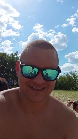 The video opens with a close-up of a man wearing sunglasses and smiling. The sunglasses reflect the sky with clouds, and what appear to be two guitars. The man is shirtless and appears to be on a beach. In the background, there are trees and a body of water. As the video progresses, the camera pans out to reveal more of the beach scene. There are other people visible on the beach, some sunbathing and some in the water. There are also some flip-flops on the sand. The man in the foreground continues to smile and look at the camera. The sky is blue with scattered white clouds, and the sun is shining. The overall impression is a sunny day at the beach.