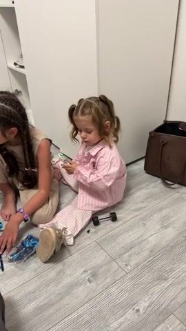 A young girl with light brown hair is sitting on a gray wood floor in a light room next to another person who is mostly out of the frame on the left. The girl is wearing pink checkered pajamas with a matching button up shirt. Her hair is in pigtails with ribbons. She is holding a black cylindrical object with a silver inner cylinder and looking at it. She turns to her left, then to her right. A set of wheels and axles is on the floor in front of her.