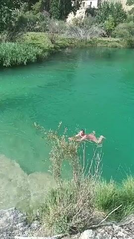 A person in red swimwear floats on their back in clear turquoise water. The person is positioned in the center of the frame, with their arms outstretched. Green reeds and grasses grow along the rocky bank in the foreground. In the background, a wooded hillside with a building is visible. The water appears calm with some ripples and small bubbles. The lighting suggests a sunny day.