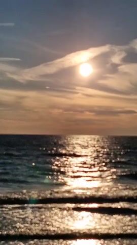 A serene ocean sunset with the sun peeking through clouds, casting a shimmering path of light across the water. The waves are gentle, and the sky is a blend of orange, yellow, and blue hues.
