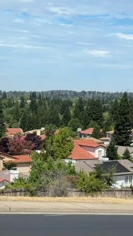 The video shows a panoramic view of a suburban neighborhood with houses nestled among trees. The houses are of various sizes and architectural styles, with some featuring red tile roofs and others with solar panels. The landscape is lush and green, with numerous trees of different species, including tall evergreen trees. The sky is partly cloudy, with white clouds scattered across a blue sky. In the distance, hills are visible, covered in more trees. The foreground shows a paved surface, likely a road or sidewalk, with dry grass and vegetation along the edge. The overall impression is a peaceful and well-established residential area.