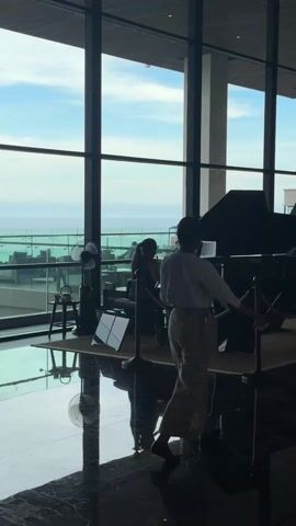 A long shot indoors depicts a woman playing a piano in front of a large floor-to-ceiling window, while another woman walks in the opposite direction in the foreground. The pianist, seen from the back, sits at a black grand piano. A music stand with sheet music is in front of her. A small black fan stands nearby. A rope barrier surrounds the small platform where the piano is placed. The window offers a view of the ocean. The floor is a shiny black marble. The second woman, facing partially to the right, is wearing a white top and light-colored patterned pants.