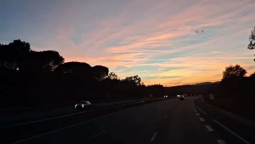 The video shows a view from inside a vehicle driving on a highway at sunset. The sky is a gradient of colors, from orange and yellow near the horizon to pink and blue higher up. Wispy clouds stretch across the sky. The road ahead is visible, with a few cars driving in the same direction. The sides of the road are lined with dark silhouettes of trees and hills. The lighting suggests it is dusk, with the sun having recently set. The overall atmosphere is calm and serene.