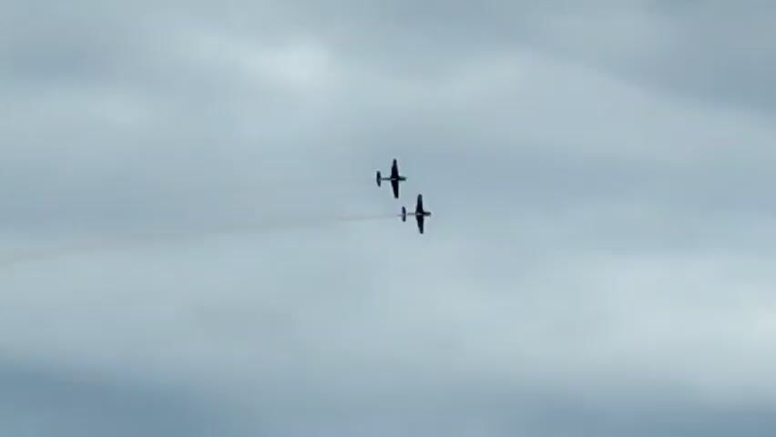 Two dark-colored propeller planes fly in tandem across a cloudy sky, leaving thin trails of smoke behind them. The planes appear to be at a slight angle to each other, possibly performing a synchronized maneuver. The sky is overcast with shades of gray and white clouds. The image is somewhat grainy and the planes are distant.