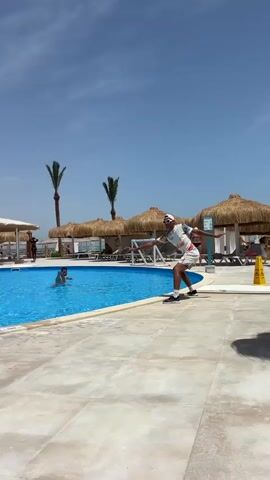 The video shows an outdoor scene at a resort swimming pool. It is a sunny day with a clear blue sky and a few palm trees are visible in the background. Several people are in the pool, and another man is standing at the edge of the pool, taking photos with his phone. The pool is surrounded by a tiled deck, and there are several thatched-roof structures nearby, likely providing shade for guests. The overall atmosphere appears relaxed and vacation-like.