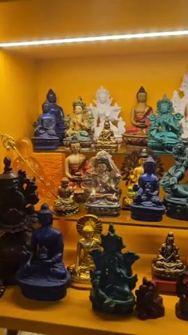 This is a medium shot video taken in a dimly lit room with yellow lighting. The scene displays a collection of small statues of Buddhist figures arranged on shelves. The statues are made of various materials and colors, including blue, green, white, gold, and dark brown. Some of the statues depict Buddha in different poses, while others appear to be deities. The statues are detailed and intricate. The background is a solid yellow color. The video is shaky, suggesting it was filmed handheld. There are no people visible in the video.