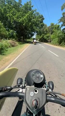 This is a first-person perspective video recorded from the handlebars of a motorcycle. The motorcycle has a surfboard strapped to its side. The road ahead is paved and appears to be a rural or semi-rural area. On either side of the road are lush green trees and vegetation, with some glimpses of buildings and power lines. The sky is a clear blue, suggesting fair weather. The time of day appears to be midday, given the bright sunlight and shadows.

In the distance, there are other vehicles, including what looks like a tuk-tuk and possibly another motorcycle. The rider is moving forward, indicating travel. The overall mood is one of leisurely exploration, possibly on the way to or from a beach.

The visible text on the motorcycle's instrument cluster appears to be part of the speedometer and other gauges, but the specific markings are not legible. The location context provided, Bodagama, Sri Lanka, further supports the tropical, sunny setting. The imagery suggests a warm, vibrant environment.