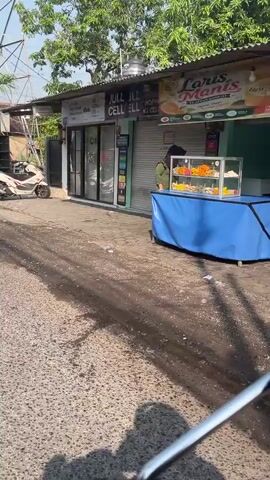 The video shows a street scene, likely in a commercial area, from the perspective of someone riding a motorcycle. In the foreground, the handlebars and a portion of the motorcycle's front are visible, suggesting a first-person point of view. The road surface appears to be asphalt, with some debris and wet patches. To the left of the road, a white scooter is parked.

The main focus of the scene is a row of storefronts. The first storefront, labeled "Jull Cell," is partially visible on the right. Next to it, a shop with a glass facade and signs displaying mobile phone plans and services is prominent. A sign above this shop reads "Jull Cell." The interior of this shop is visible through the glass, and it appears to be a mobile phone store, with displays and possibly staff inside. Further to the left, another storefront with a closed shutter is seen. Above the shops, there is a canopy or awning structure, and trees are visible in the background, suggesting an outdoor setting with vegetation. The lighting indicates it is daytime, likely sunny. The overall atmosphere appears to be that of a typical urban or semi-urban commercial street.