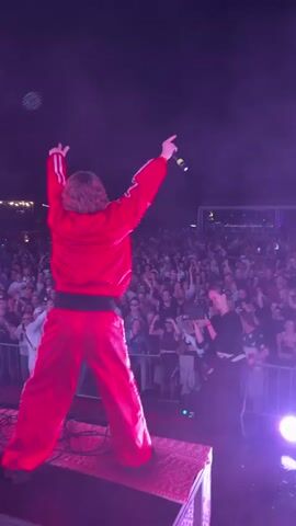 A person wearing a bright red tracksuit with white stripes on the sleeves and legs stands on a platform on a stage. The person has long, wavy brown hair and is facing away from the camera, looking out at a large crowd. They are holding a microphone in their right hand and have their left arm raised in the air, with their fist clenched. Their right arm is also raised, with their fist clenched. The stage is lit with purple and pink lights, and there is a lot of fog or haze in the air. The crowd is visible behind a metal barrier, and many people are holding up their phones to take pictures or videos. The overall atmosphere appears to be that of a live concert or performance.