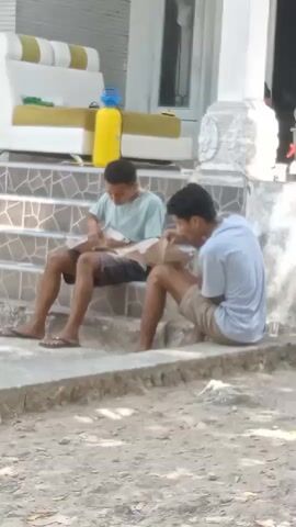 TWO YOUNG MEN SIT ON A STAIRCASE OUTSIDE A BUILDING. THEY ARE BOTH WEARING T-SHIRTS AND SHORTS. THEY ARE EACH HOLDING A BROWN PAPER BAG AND APPEAR TO BE EATING OR DRINKING FROM THEM. THE MAN ON THE LEFT IS WEARING A LIGHT BLUE T-SHIRT AND DARK SHORTS. THE MAN ON THE RIGHT IS WEARING A LIGHT BLUE T-SHIRT AND KHAKI SHORTS. THE BACKGROUND SHOWS A PORCH WITH A WHITE SOFA AND PILLARS. THE GROUND IS DIRT AND GRAVEL.