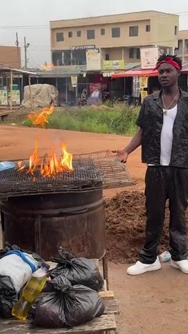 The video shows a dark-skinned man standing next to a fire on top of a metal barrel. He is wearing a white t-shirt, black jacket, black pants, and white shoes. He also has a red bandana tied around his head. The fire is large and brightly colored. To the left of the barrel is a wooden table with several black trash bags and a bottle of oil. In the background, there are several buildings and a dirt road. A car drives behind the man to the left, and another man walks past him as well. It looks like a street-side grill or food stall set up.