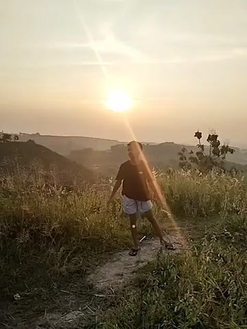 A young boy walks along a narrow dirt path through a grassy, hilly landscape during golden hour, suggesting either sunrise or sunset. The bright sun is low in the sky, positioned behind him, creating a strong lens flare and illuminating the scene with a warm, soft light. The boy is wearing a black t-shirt with a white logo on the front, light-colored plaid or striped shorts, and flip-flops. Tall, dry-looking grass and sparse bushes line the path. In the initial moments, the boy looks upwards, seemingly interacting with the sunlight, before continuing to walk forward, glancing around at his surroundings. Distant hills and an expansive, clear sky are visible in the background, indicating an open, natural setting in a location such as Jambeyan, Indonesia.