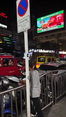 The video shows a busy street scene in Thailand at night. There are many vehicles including tuk-tuks and cars moving slowly through traffic. Several motorcycles are parked along a metal fence. People are walking on the sidewalks and sitting at outdoor tables. There are multiple bright signs and billboards advertising restaurants and products, including a large Pizza Hut billboard. The overall impression is of a vibrant, crowded urban environment at night.