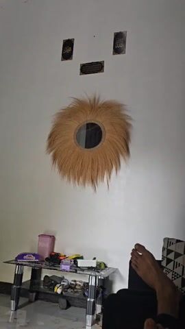 A mirror is on the wall. It is round and has a sunburst of straw around it. The straw is long and golden brown. In the background are some pictures on the wall and a shelf with some items on it. A person's foot is visible on the right side of the frame, resting on what appears to be a couch.