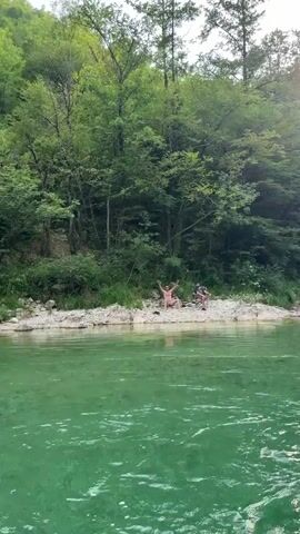 The media file shows a serene outdoor scene by a clear, turquoise river. Two men are the primary subjects, both appearing to be in their 20s or 30s. The man on the left is seated on a rocky bank, wearing light-colored shorts and no shirt. He has sunglasses on and his arms are raised in a celebratory gesture, with a phone or camera in his right hand. The man on the right is sitting in a folding chair, wearing a dark t-shirt and shorts. He is looking towards the camera and appears to be pointing or gesturing.

The setting is a natural environment with lush green trees and dense foliage forming a backdrop behind the men and the riverbank. The river itself is the foreground, its surface calm and reflecting the surrounding greenery. The riverbank is composed of pebbles and rocks. The overall atmosphere suggests a leisurely day spent outdoors, possibly a break during a trip or an outing to enjoy nature. The lighting suggests it's daytime, likely mid-morning or afternoon, with no strong indication of specific weather conditions beyond being clear enough for outdoor activities. There is no visible text in the media file. The context provided places this scene in Zagorice, Bosnia and Herzegovina.