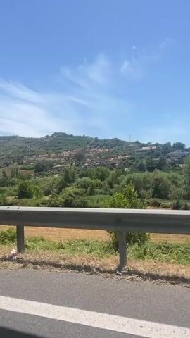The video shows a roadside view with a metal guardrail in the foreground and a hilly landscape with trees and houses in the background under a clear blue sky with some white clouds. A white sign with red, blue, and black lettering is visible on the left. A light blue car is seen on the right. The camera is moving, suggesting it's from a vehicle. The scene appears to be a rural or semi-rural area.