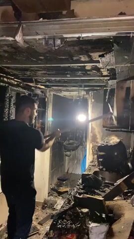 The video shows a person holding a long tool and surveying a room that appears to have been damaged by fire. The ceiling and walls are blackened and charred, with debris scattered on the floor. The person is wearing a black t-shirt and dark pants. The room appears to be a kitchen or bathroom, with remnants of appliances and fixtures visible. The person is examining the damage, possibly in the process of cleanup or assessment.