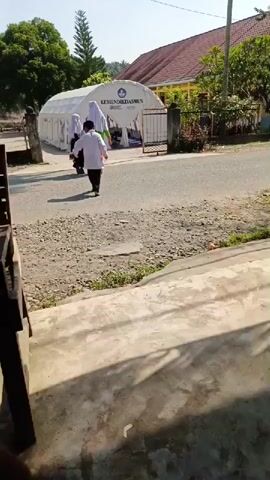 The scene is a stationary shot from what appears to be a porch or entryway, with a partial view of a wooden structure to the left. The camera is focused on a street leading to a white tent structure in the middle ground, labeled "KEMENDIKDASMEN." Three figures are walking towards the tent. Two individuals, likely girls, are wearing traditional school uniforms, one in black with a white hijab and the other in black with a white hijab and an orange skirt. A third, smaller figure, likely a boy, is wearing a white shirt and dark pants. In the background, there is a building with a red tile roof and green foliage. The day appears bright and sunny.