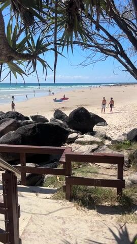The video presents a view of a sun-drenched beach from behind a short, wooden railing. In the foreground, there's a collection of large, dark boulders scattered on the sandy shore, some interspersed with lighter-colored rocks. A person is seen lying on the sand between the boulders.

The beach extends towards the ocean, and several people are visible engaging in various activities. Some are walking along the shore, while others are in the water. A red surfboard lies on the sand. The ocean stretches to the horizon under a clear blue sky with a few scattered clouds.  Palm trees are visible in the upper left and right corner, framing the shot, their fronds adding a tropical touch to the scene.
