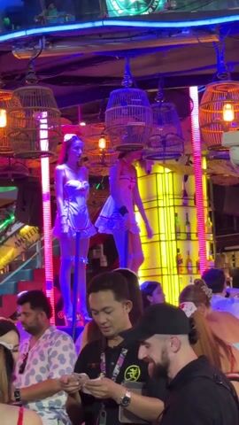 The scene unfolds inside a bustling, brightly lit bar or club with a highly decorated interior. The space is crammed with people, most of whom seem to be engaging in casual conversations or observing the spectacle above. Two young women, dressed in short, light-colored dresses, high heels, and headbands, dance on elevated platforms, each equipped with a pole. The interior decorations are a wild mix, including illuminated birdcages hanging from the ceiling, neon signage, and shelves stocked with liquor bottles. In the foreground, a group of individuals is clustered around a table, seemingly engrossed in a mobile phone. The overall atmosphere suggests a lively and somewhat provocative social gathering.